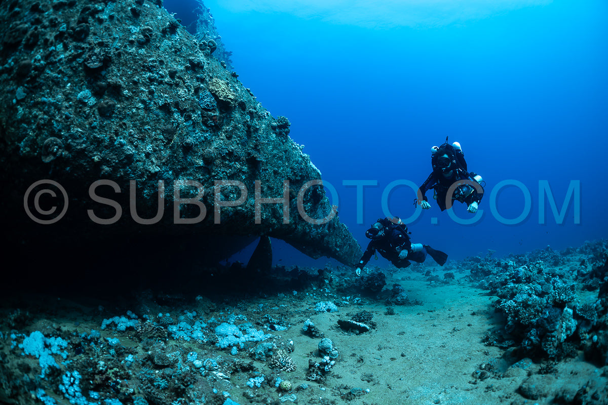 Two divers using rebreathers swim past a large rock formation in a clear blue sea. The divers are wearing full-face masks and have their dive lights on.