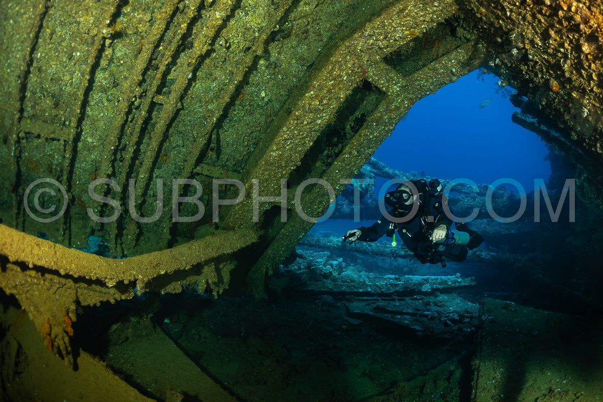 A diver wearing a rebreather swims through a large opening in a shipwreck. The interior of the ship is dark- but the surrounding water is clear and blue.