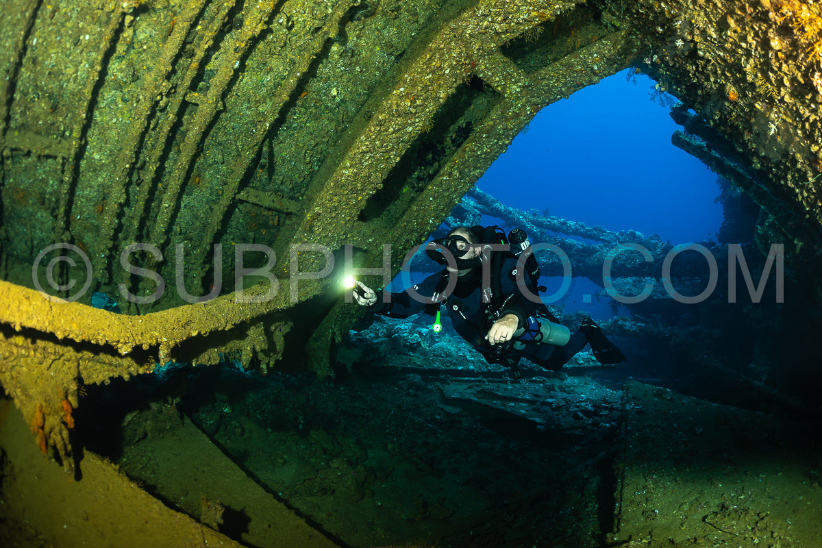 A diver with a rebreather swims through a large opening in a shipwreck- illuminating the dark interior with a flashlight.