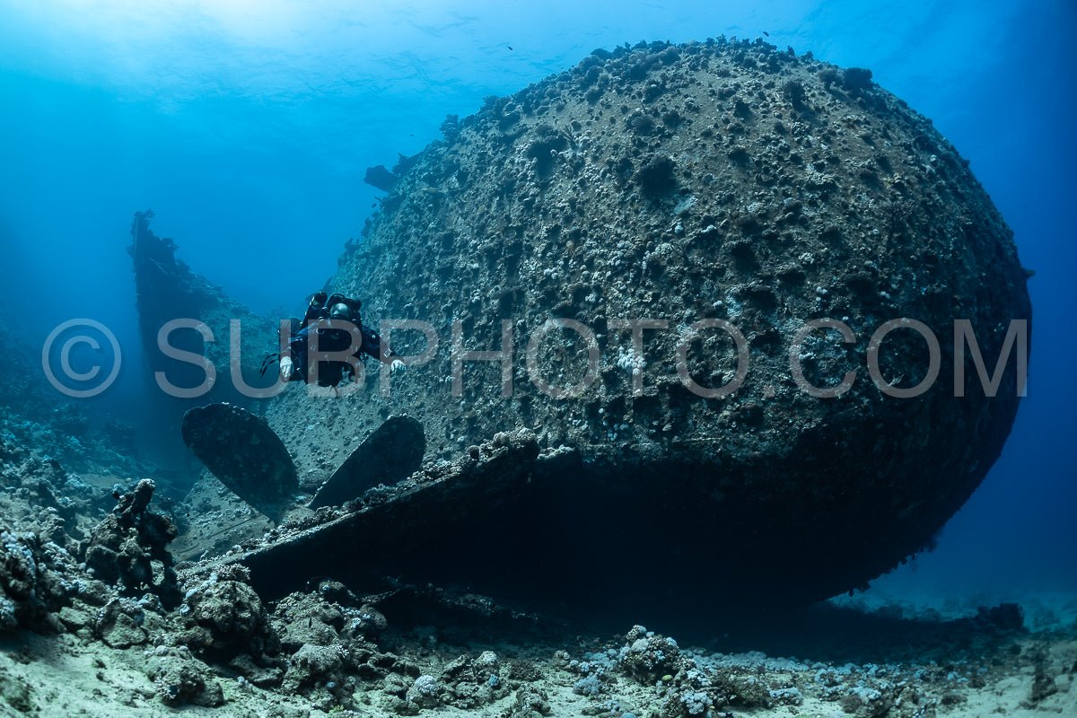 A diver in full scuba gear swims past a large wreck on the seabed. The diver is using a rebreather and is silhouetted against the blue water.