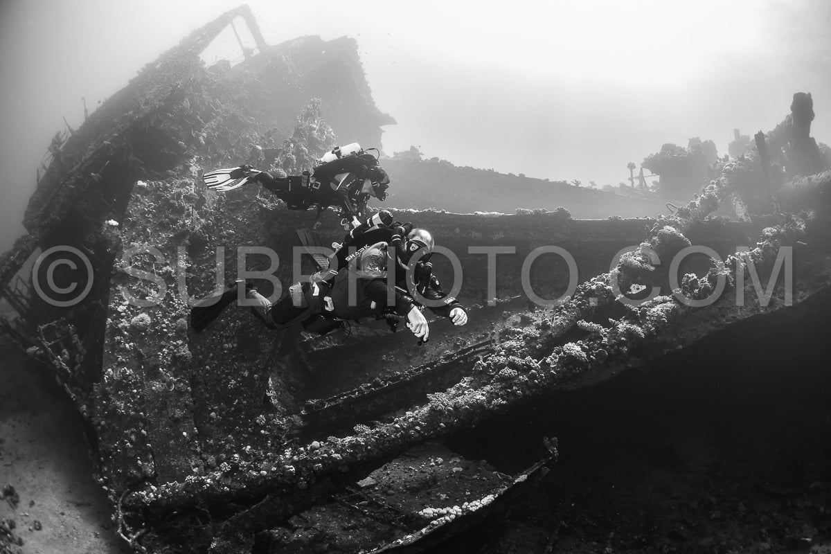 Two divers- wearing scuba gear and using a rebreather- are exploring a shipwreck in the Red Sea. The shipwreck is partially covered in marine life.