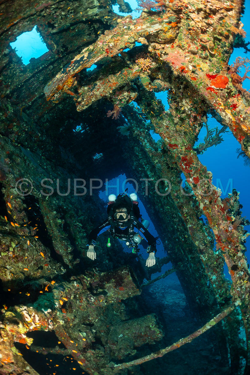 A diver wearing a rebreather swims through the interior of a shipwreck. The shipwreck is covered in coral and other marine life. The diver is surrounded by a school of fish.