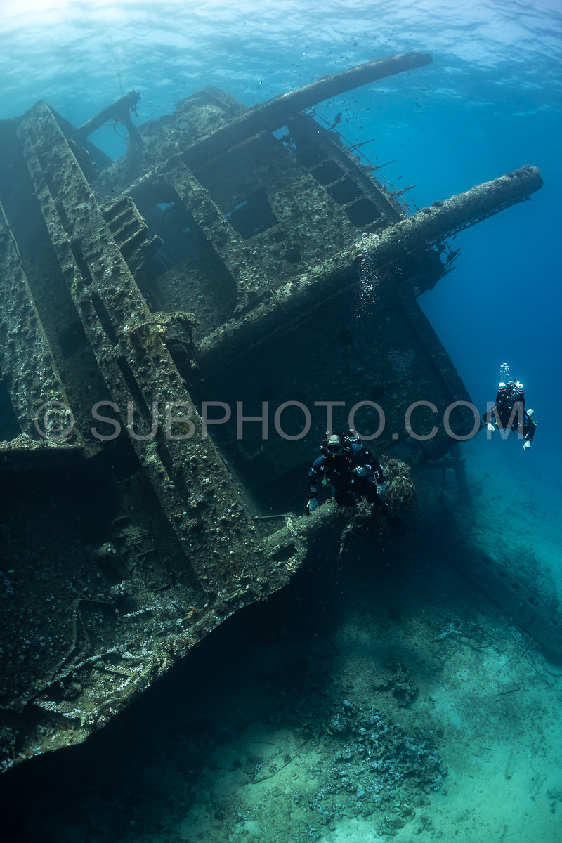 Two divers in full scuba gear are exploring a large shipwreck in the Red Sea. The wreck is covered in algae and coral- and the water is clear and blue.