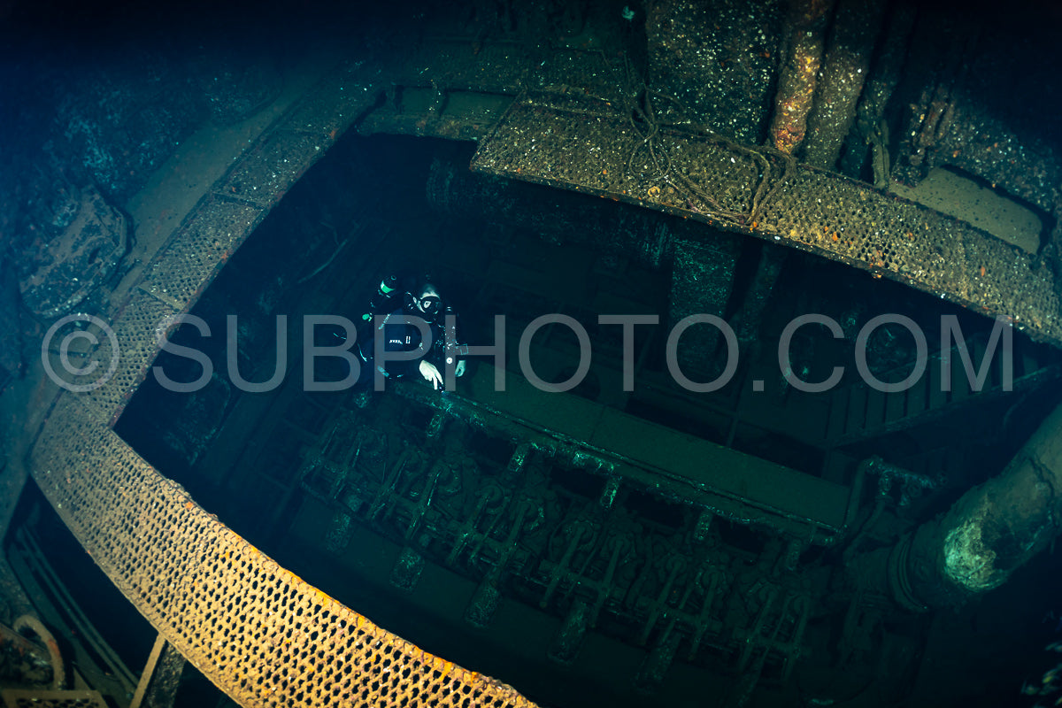 A diver is seen swimming inside the open hold of a sunken ship. The diver is using a rebreather- allowing for extended exploration. The ship's interior is dark and covered in marine life.