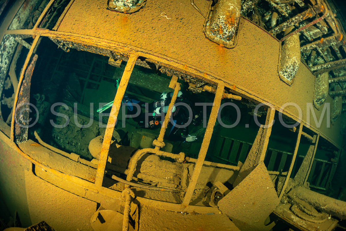 A diver wearing a rebreather is inside the rusted interior of a shipwreck in the Red Sea. The diver is illuminated by a light- casting a greenish glow on the surrounding metal.
