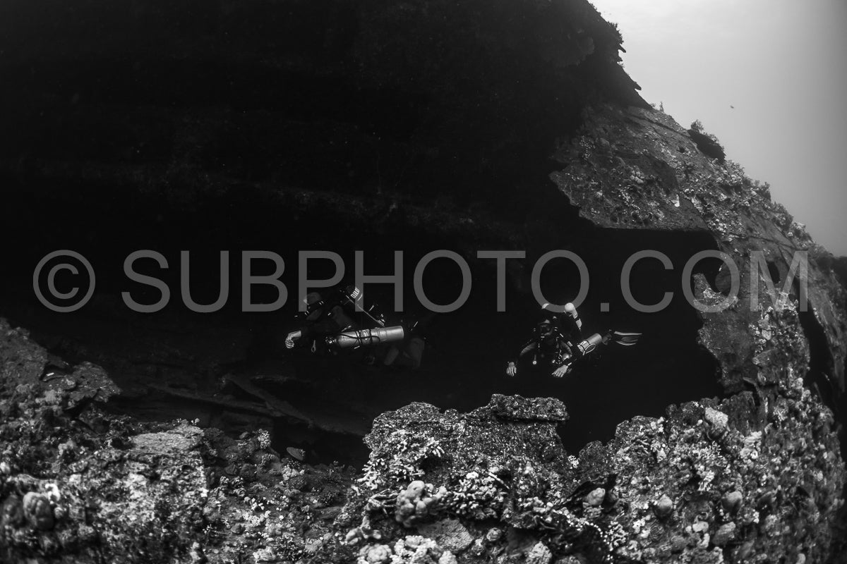 Two divers are seen navigating a dark- underwater passage in a shipwreck. The divers are using rebreathers and are silhouetted against the darkness.