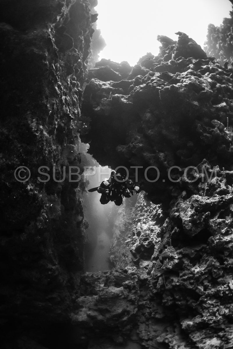A diver with a rebreather swims through a narrow crevice in a coral reef in the Red Sea. Sunlight streams in from above- illuminating the diver's silhouette.