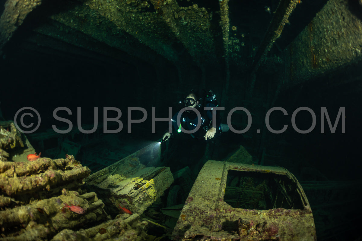 A diver- wearing a rebreather and carrying a flashlight- navigates the dark and overgrown interior of a sunken ship.
