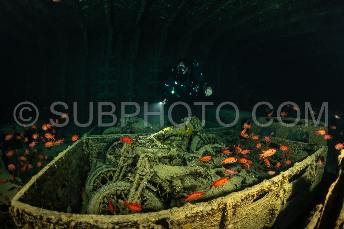 A diver wearing a rebreather swims through a dark- murky wreck in the Red Sea. Their flashlight illuminates a motorcycle lying on the ocean floor. Schools of small- orange fish swim around the wreck.