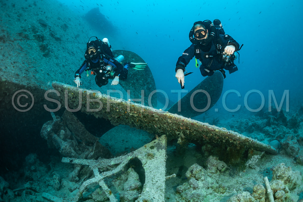Two scuba divers- using rebreathers- swim past a large shipwreck in the Red Sea. The divers are positioned above the wreck- their fins extended behind them.