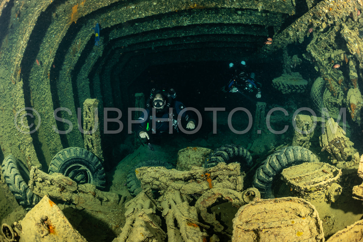 A diver- wearing a rebreather- is exploring the interior of a shipwreck in the Red Sea. The wreck is covered in algae and barnacles- and the interior is dark and mysterious.
