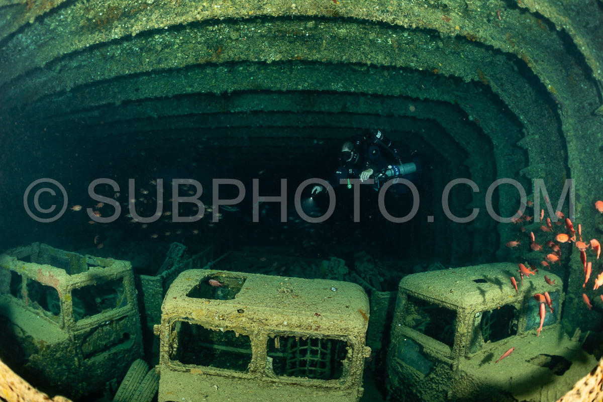 A diver wearing a rebreather swims through a large open space inside a shipwreck. The wreck is covered in algae- and the diver's light illuminates the dark interior.