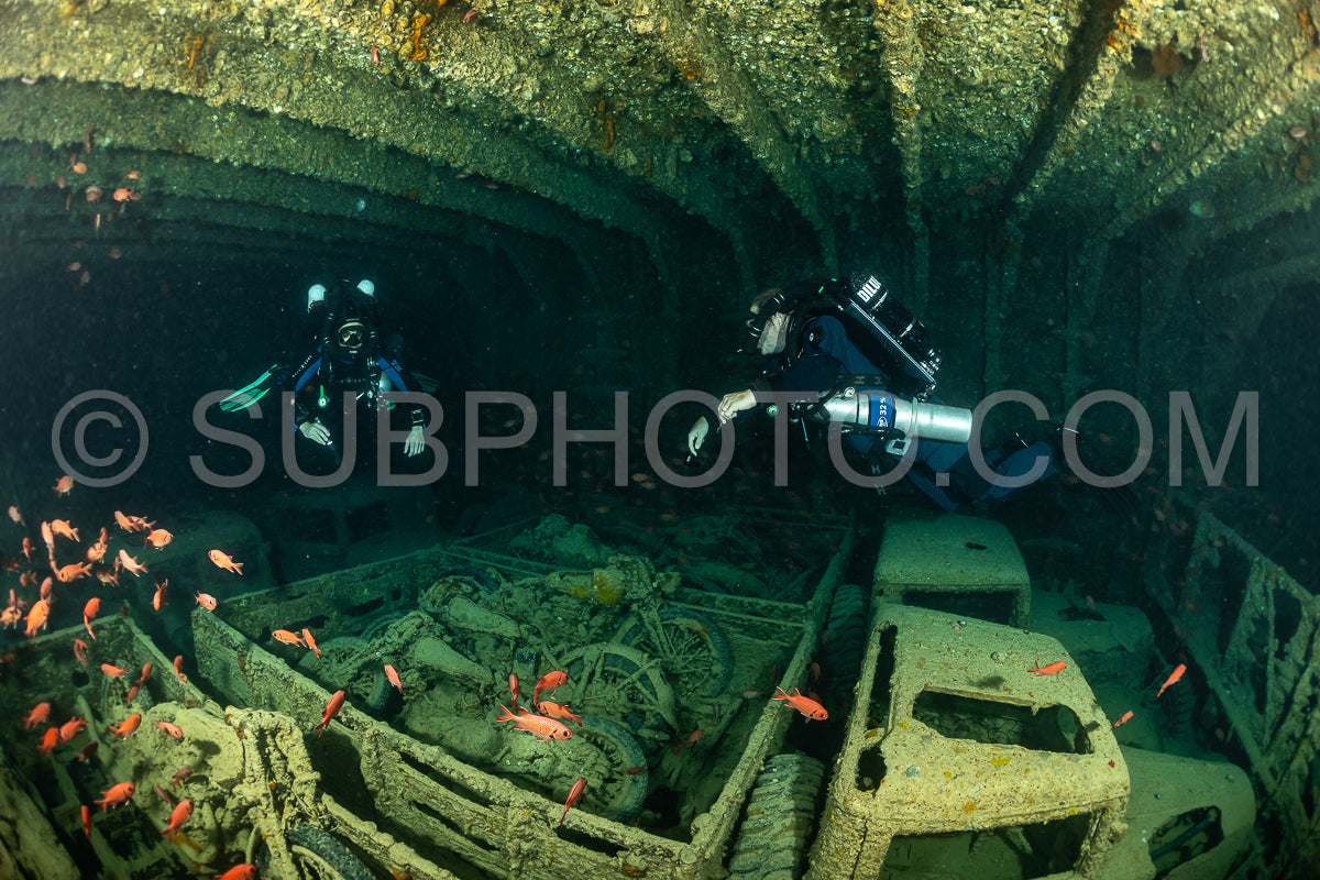 Two divers- equipped with rebreathers- swim through the cargo hold of a sunken ship. The hold is filled with vehicles and debris- covered in marine life. Red fish swim around the divers.