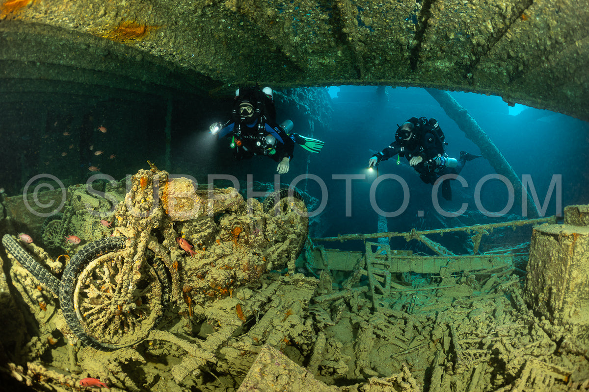 Two scuba divers with lights explore the interior of a shipwreck in the Red Sea. The wreck is overgrown with coral and other marine life.