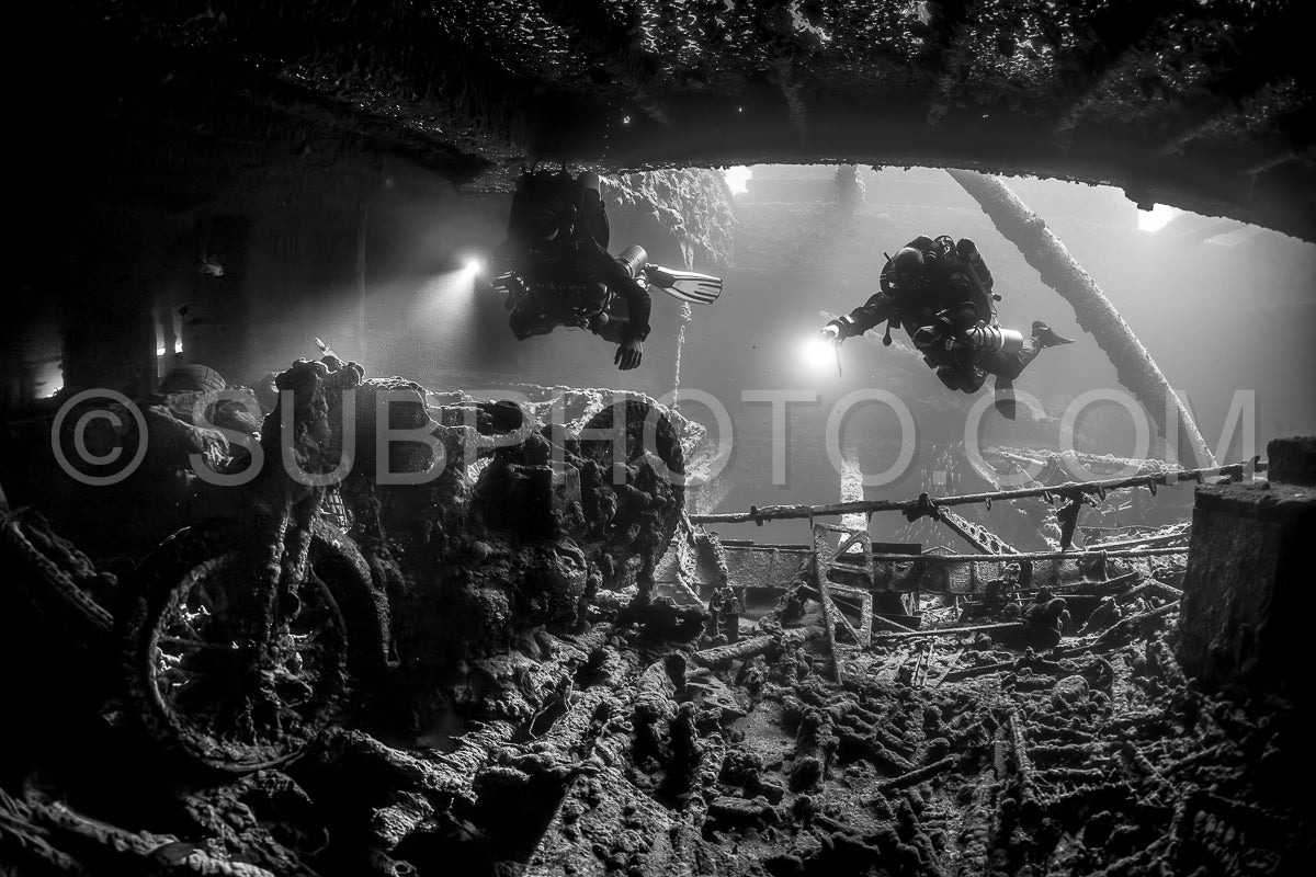 Two divers- using rebreathers- explore a shipwreck in the Red Sea. Their beams illuminate the dark interior- revealing a large wheel and other debris.