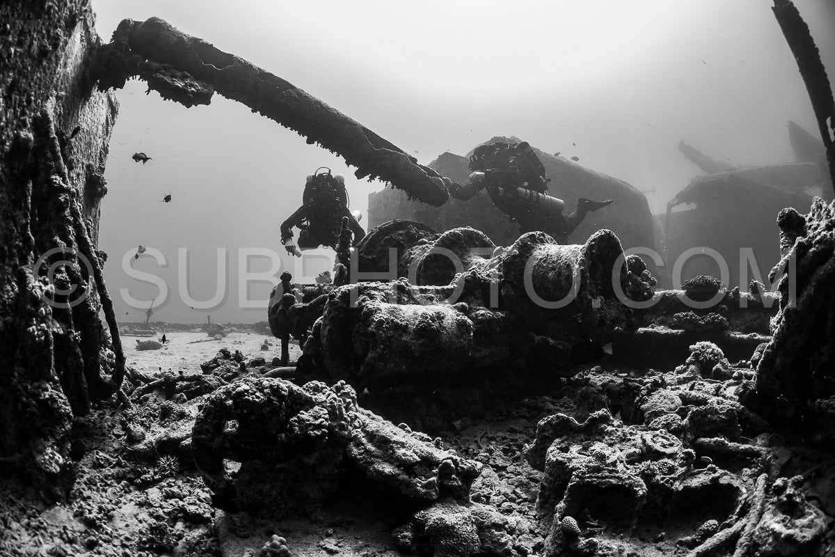 A diver with a rebreather is exploring the wreck of a ship in the Red Sea. The diver is silhouetted against the sunlight filtering through the water- and the wreck is covered in marine life.