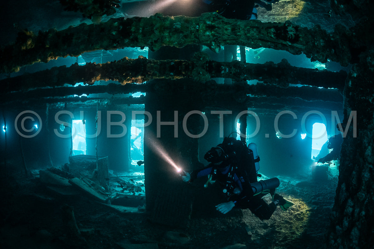 A lone diver- equipped with a rebreather- navigates the darkened interior of a submerged shipwreck.