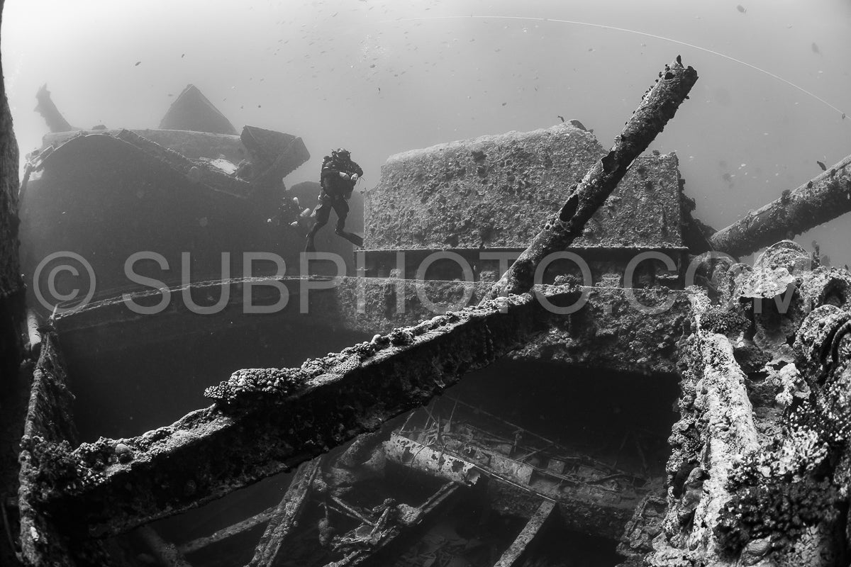 A diver- wearing a closed-circuit rebreather- swims through a sunken ship in the Red Sea. The ship is heavily encrusted with marine life and the diver's form is silhouetted against the dim light.