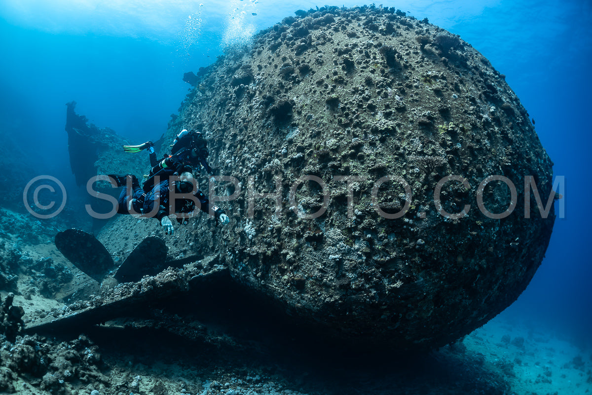 Two divers are swimming near a large- round shipwreck in the Red Sea. The divers are using rebreathers and are exploring the wreck.