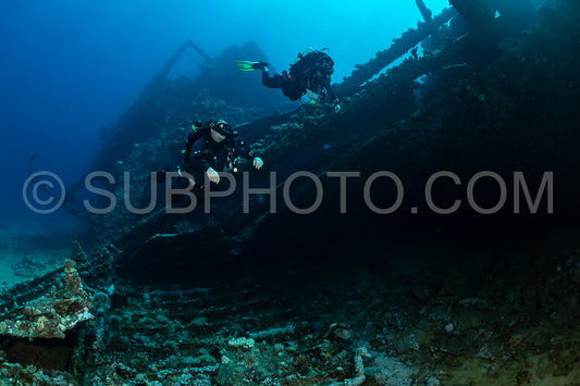 Two divers wearing scuba gear swim along the side of a sunken shipwreck in the Red Sea. They are using rebreathers to breathe underwater. The wreck is overgrown with coral and marine life.