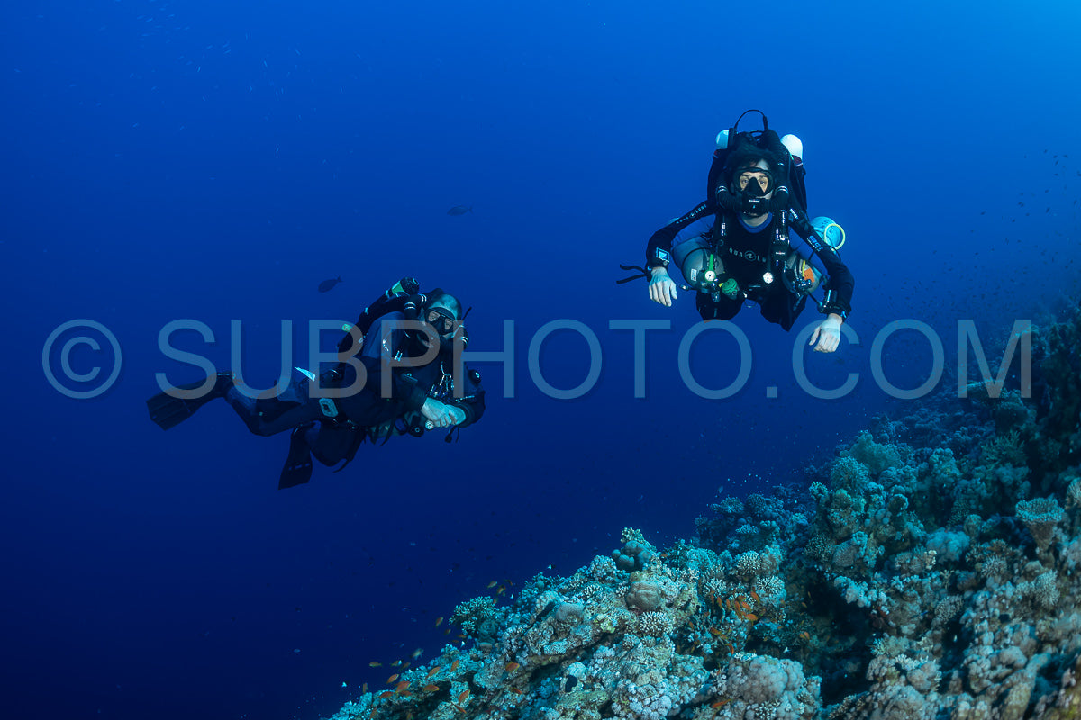 Two divers- wearing rebreathers- are swimming in the Red Sea. The water is clear and blue- and the divers are silhouetted against the light. There is a coral reef in the background.