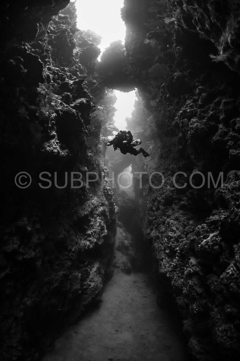 A diver swims through a dark- narrow channel in the Red Sea. The diver is using a rebreather and is illuminated by a shaft of light from above.