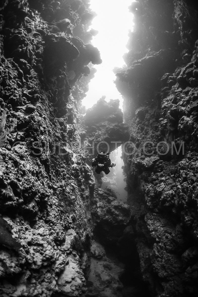 A diver using a rebreather swims through a narrow underwater canyon- sunlight streaming in from above. The walls of the canyon are covered in textured coral formations.