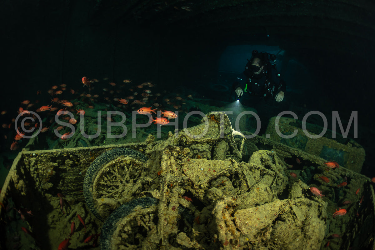 A diver wearing a rebreather explores a sunken motorcycle in the Red Sea. The motorcycle is covered in algae and coral- and the diver is using a light to illuminate the scene.