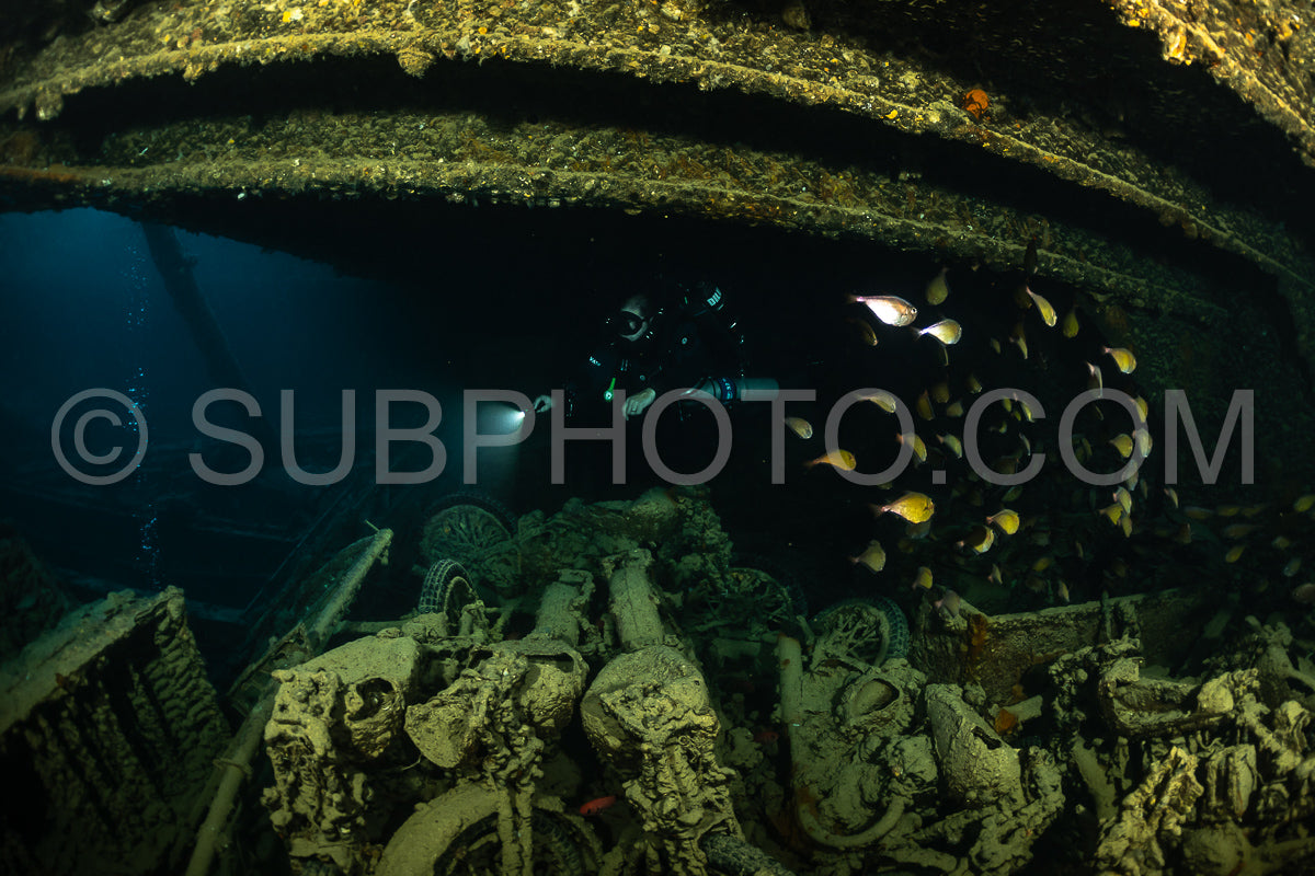 A diver wearing a rebreather is exploring a shipwreck in the Red Sea. The diver's light illuminates the wreckage and a school of fish.