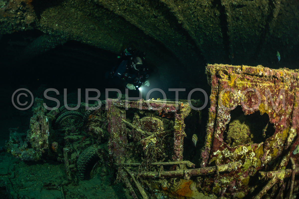 A diver using a rebreather swims through a dark underwater tunnel in the Red Sea- illuminating a rusted shipwreck with their light.