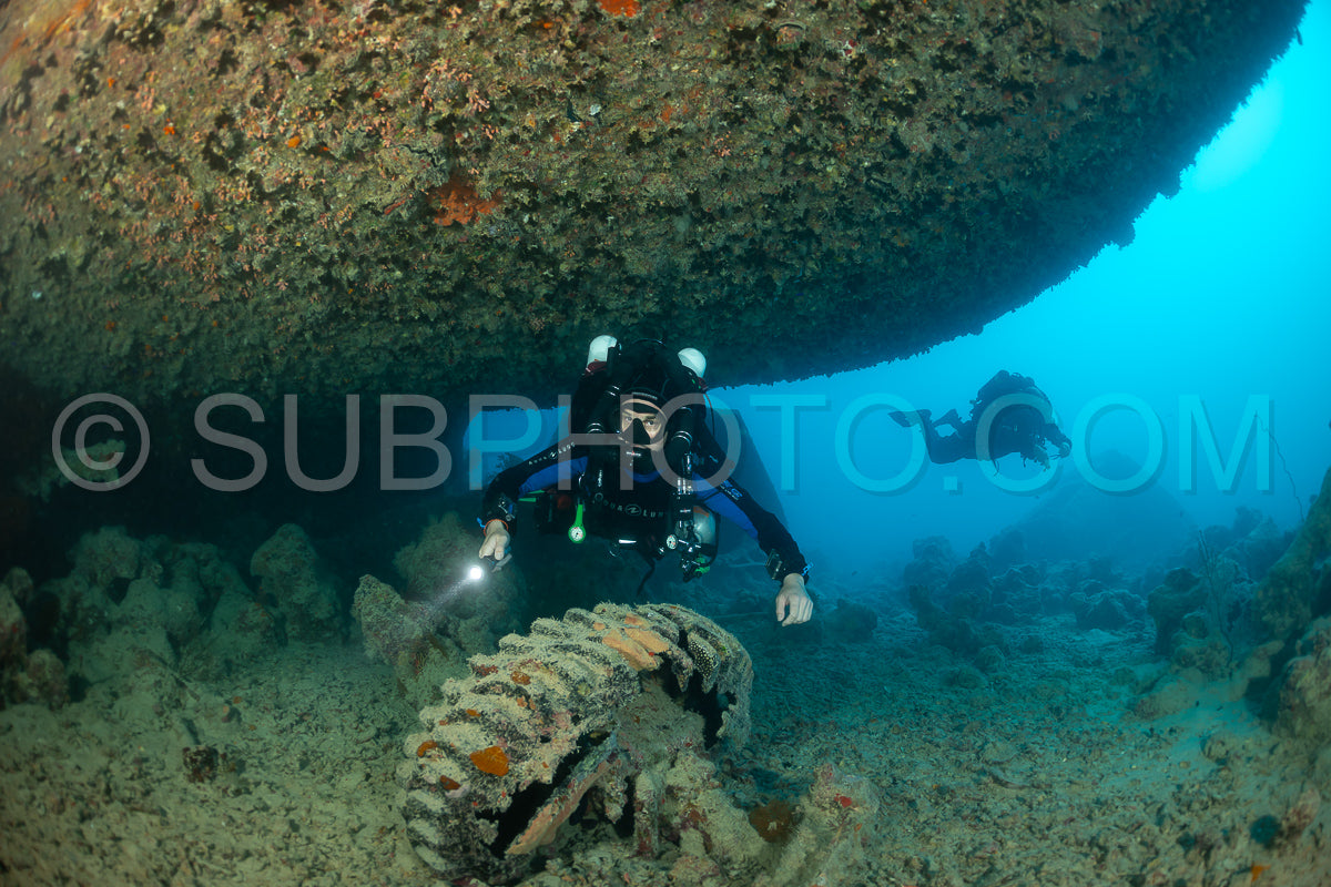 A diver wearing a rebreather swims under an overhang on a shipwreck in the Red Sea. They are using a flashlight to illuminate the surrounding area.