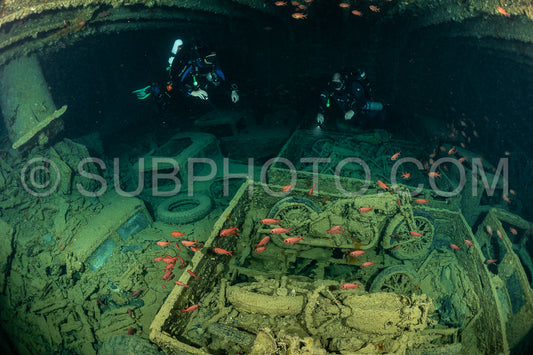 Two divers using rebreathers explore a shipwreck in the Red Sea. They are surrounded by the wreckage of vehicles- likely trucks and motorcycles- that are overgrown with marine life.