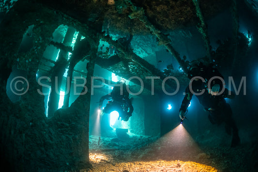 Two divers using rebreathers explore the inside of a shipwreck. The divers are illuminated by their lights- casting beams across the dark interior of the wreck.