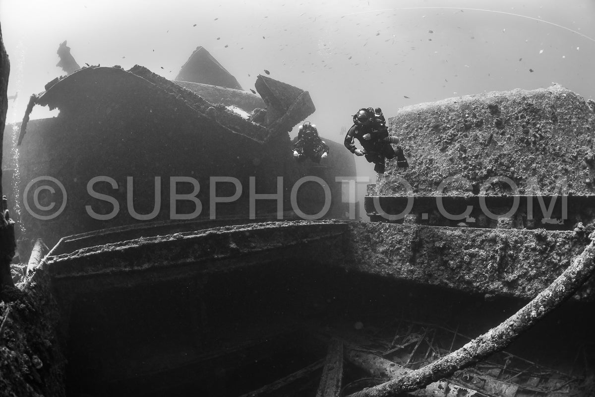 Two divers in full scuba gear explore a shipwreck covered in marine life. The divers are using rebreathers to extend their dive time. The shipwreck is located in the Red Sea.