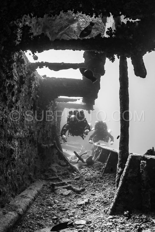 Two divers using closed-circuit rebreathers swim through the interior of a shipwreck.