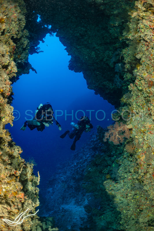 Two divers- equipped with rebreathers- swim through a narrow passageway in the Red Sea. The water is clear and blue- and the walls of the passageway are covered in colorful coral.