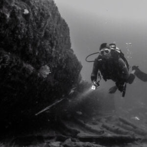 Black and white: diver with torch beside a massive encrusted hull wall, Ville de Rochefort wreck, Brittany, France