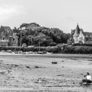 Black-and-white shellfish gatherer crouching on shore below turreted seaside manors, Le Croisic, Brittany, France