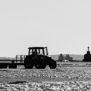 Black-and-white silhouette of tractor and trailer on tidal flat, stone cross beyond, Le Croisic, Brittany, France