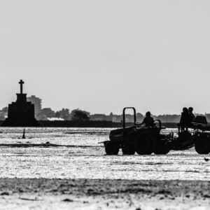 High-contrast black-and-white tractor carrying two people across flat, stone cross behind, Le Croisic, Brittany, France