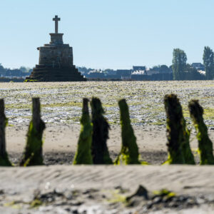 Seaweed-covered posts in foreground with stone refuge cross on low-tide sand, Le Croisic, Brittany, France