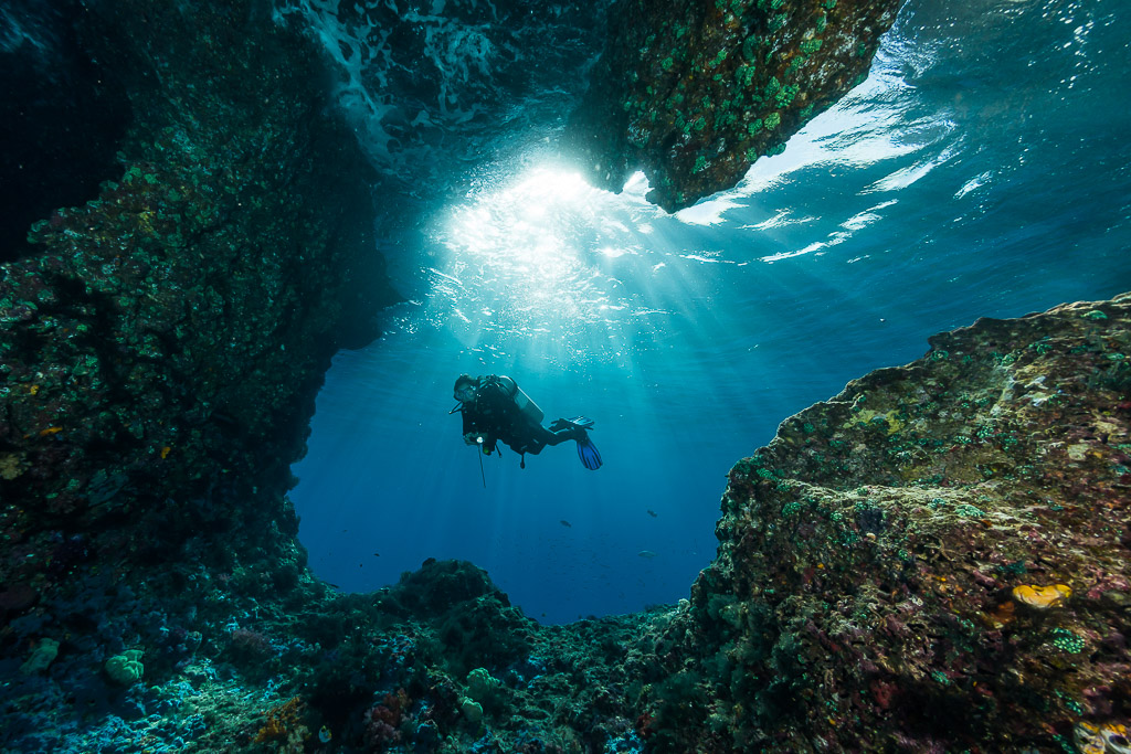 Diver silhouetted in a wide cave opening with sun rays, Misool, Raja Ampat, Indonesia