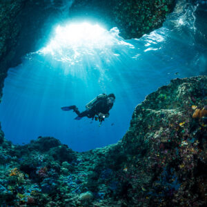 Diver swimming across a cave entrance under shafts of light, Misool, Raja Ampat, Indonesia