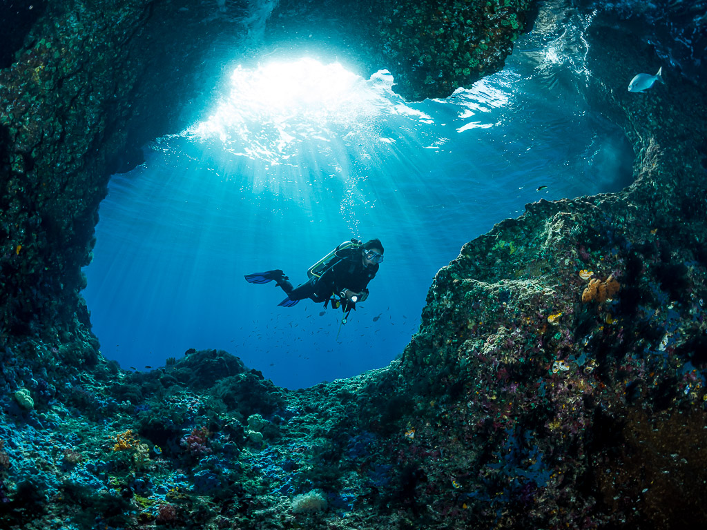 Diver swimming across a cave entrance under shafts of light, Misool, Raja Ampat, Indonesia