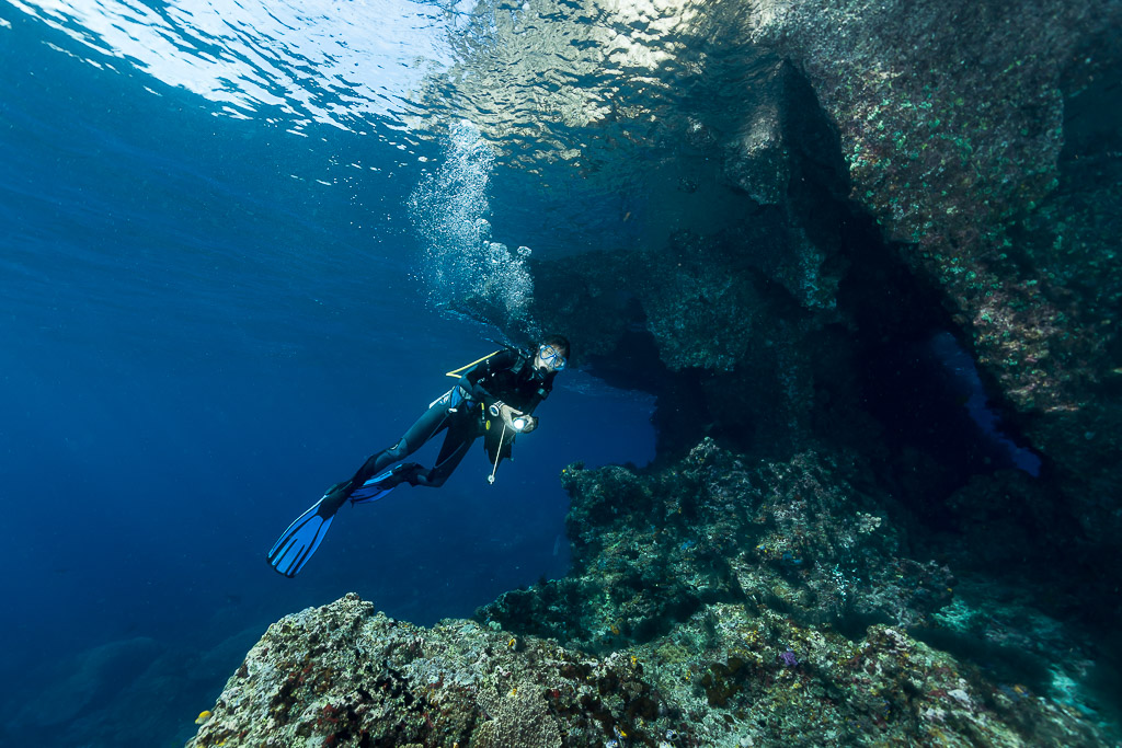 Diver ascending past a cave entrance trailing bubbles, Misool, Raja Ampat, Indonesia