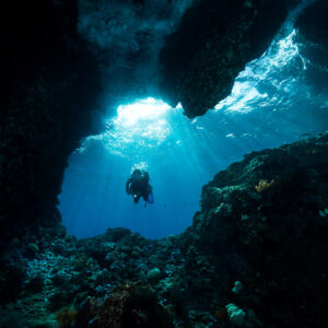 Distant diver framed in a dark cave mouth with sun rays, Misool, Raja Ampat, Indonesia
