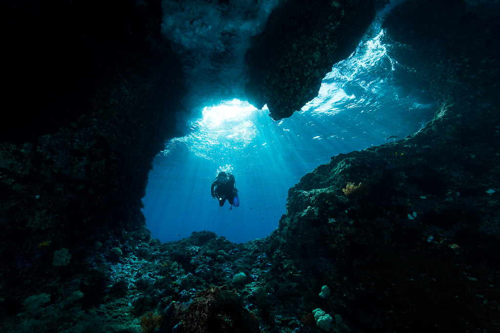 Distant diver framed in a dark cave mouth with sun rays, Misool, Raja Ampat, Indonesia