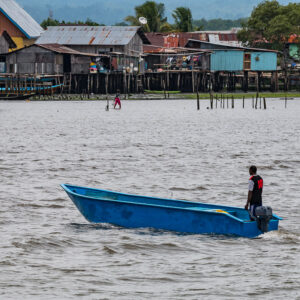 Man standing in a blue outboard boat before stilt houses, Misool, Raja Ampat, Indonesia