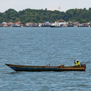 Fisherman in a small wooden canoe with distant shoreline town, Misool, Raja Ampat, Indonesia