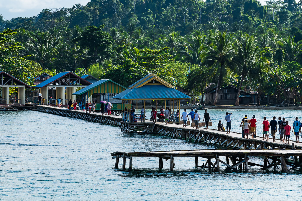 Villagers walking on a long wooden jetty to blue-roofed pavilions, Misool, Raja Ampat, Indonesia